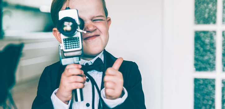 Picture: Boy with blond hair shows a thumbs up sign while shooting a video with an old analog 8 mm retro camera. The boy wears a tuxedo with a bow tie.